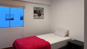A small, single-bed room with a red blanket, a white pillow, and a black nightstand. There is a window with a view of a building, and a piece of abstract art on the wall. - 3bed Apt in Miraflores - Free Wifi, Free Parking (Lima)