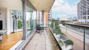 Een balkon met glazen hekwerk, een tafel en een stoel, met uitzicht op een stadsbeeld en een weg.