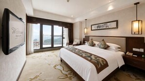 Habitación de hotel con una cama grande, un escritorio, una silla, una lámpara de escritorio, un cartel de bienvenida enmarcado y vistas al exterior.