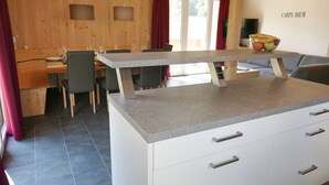 A modern kitchen with a central island, wooden walls, and a dining area with chairs and a table. - Murau 5 - Chalet Murlaub (Murau)