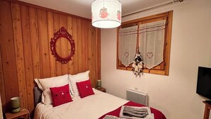 A bedroom with a wooden paneled wall, a bed with red bedding, a bedside table with a lamp, and a television on a wooden stand. - Appartement Privé Dans Chalet Proche des Cimes, Départ et Retour ski aux Pieds (Montgenèvre)