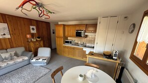 A compact living space with a kitchenette, a dining area with a round table, and a sofa with a decorative cushion. - Appartement Privé Dans Chalet Proche des Cimes, Départ et Retour ski aux Pieds (Montgenèvre)