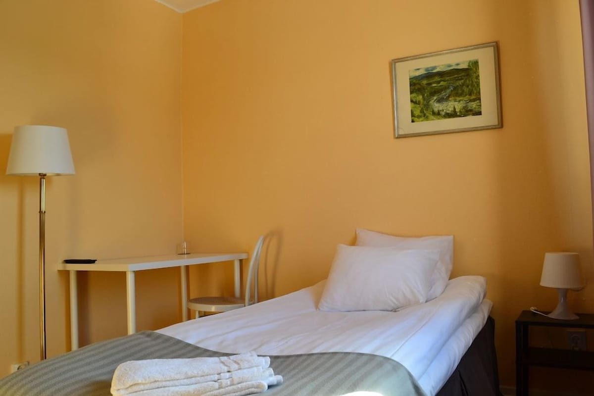 A hotel room with a bed, a desk, a chair, a lamp, and a painting on the wall.