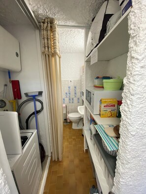 A narrow bathroom with a toilet, a shower area, and shelves with toiletries. - Cassetta 92 Marie Pierre Studio Cocooning en rdc Parking Privé Climatisé Wifi (Lumio)