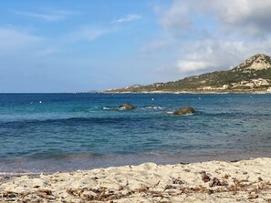 A beach with clear blue water, rocky outcrops, and a distant hill with buildings. - Cassetta 92 Marie Pierre Studio Cocooning en rdc Parking Privé Climatisé Wifi (Lumio)