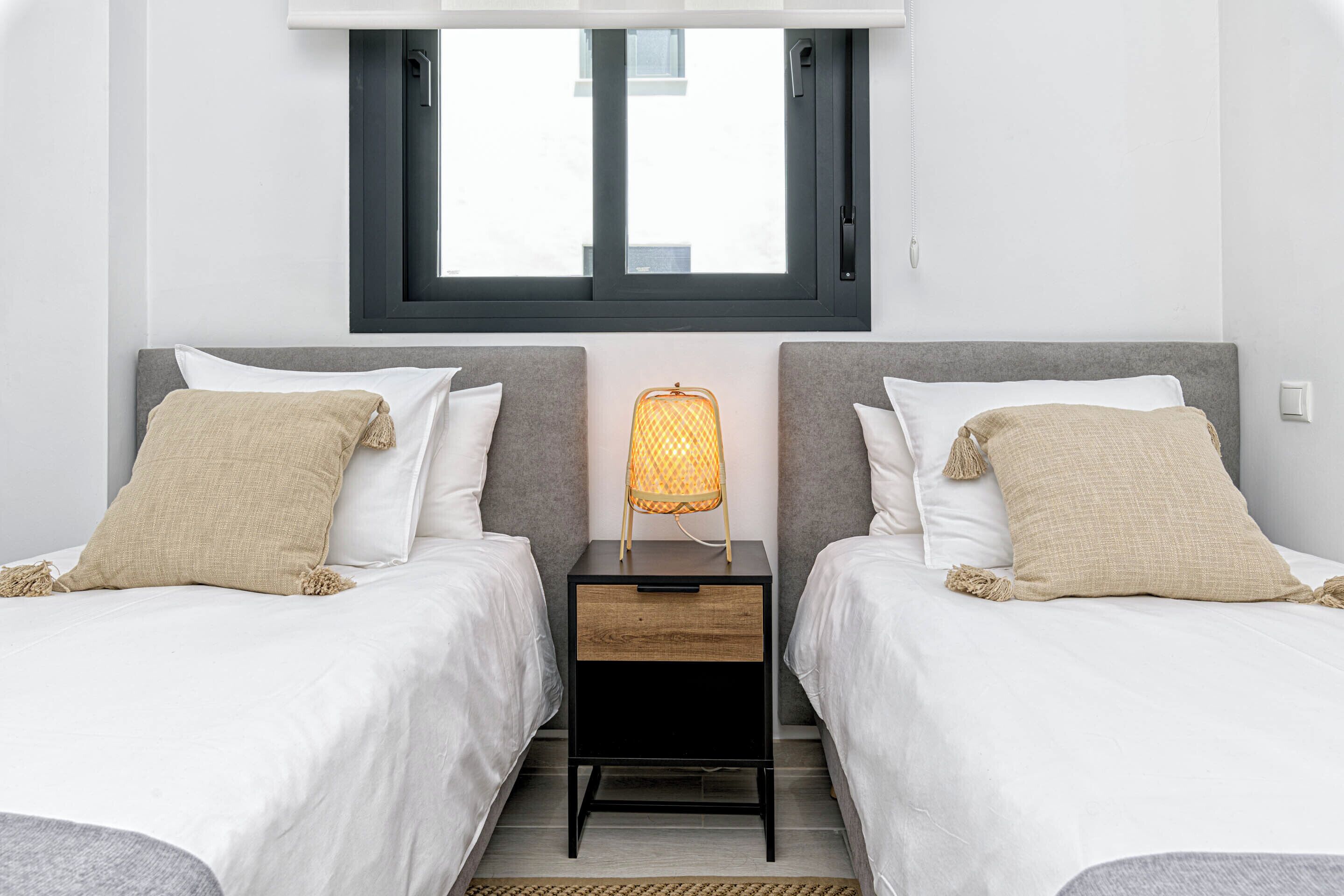 A twin room with two beds, a nightstand with a lamp, a window, and a plain wall.