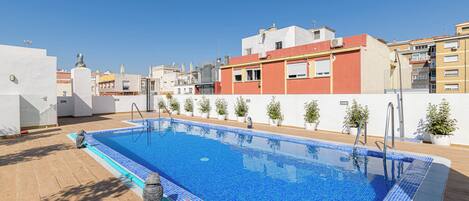 A rooftop swimming pool with a tiled edge, surrounded by potted plants and a clear blue sky.