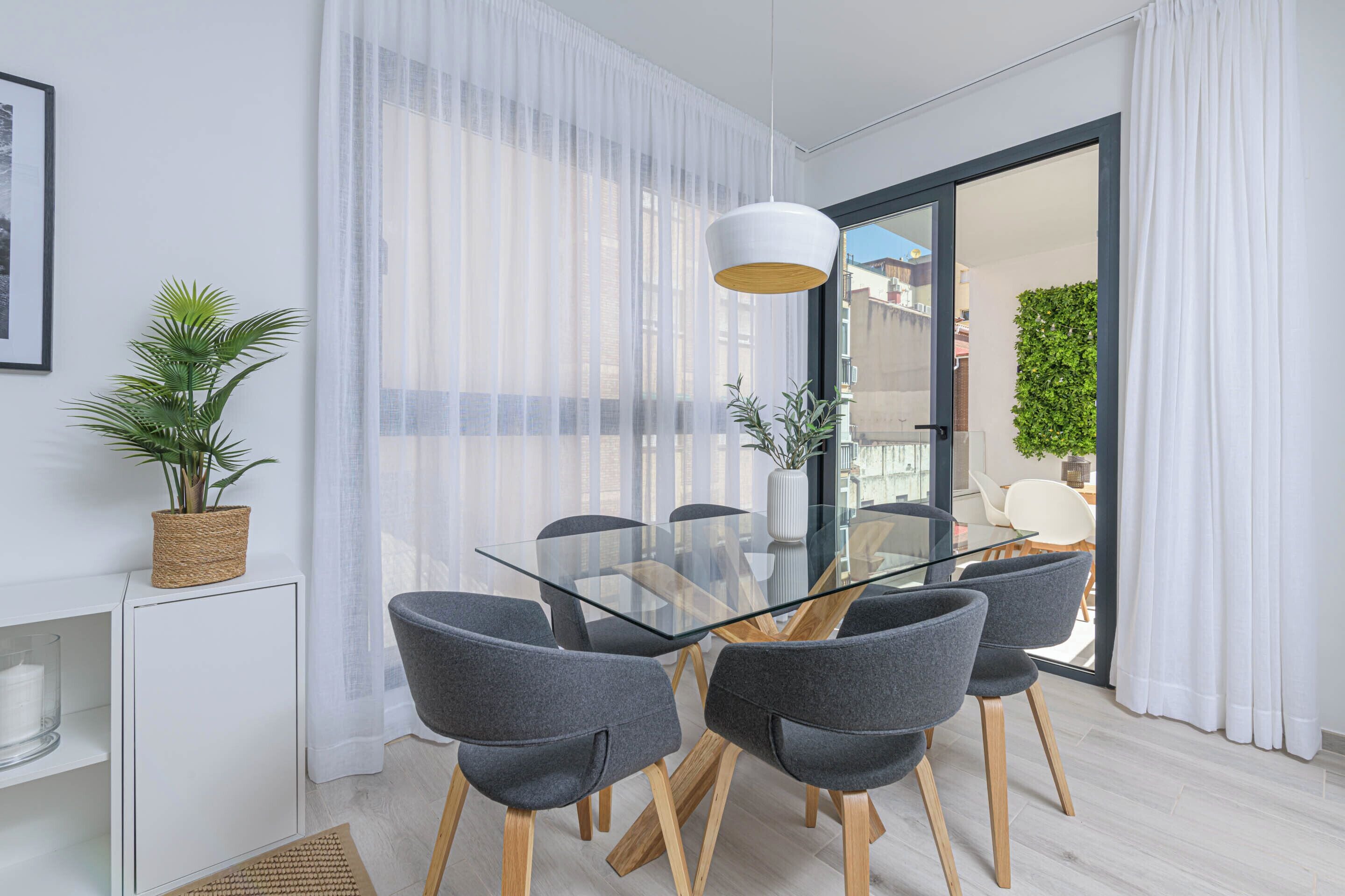 Un comedor moderno con una mesa de vidrio y sillas grises, una planta en maceta y vistas a través de una puerta de cristal.
