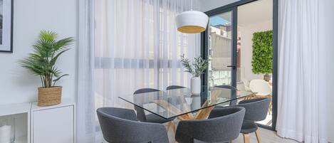Un comedor moderno con una mesa de vidrio y sillas grises, una planta en maceta y vistas a través de una puerta de cristal.