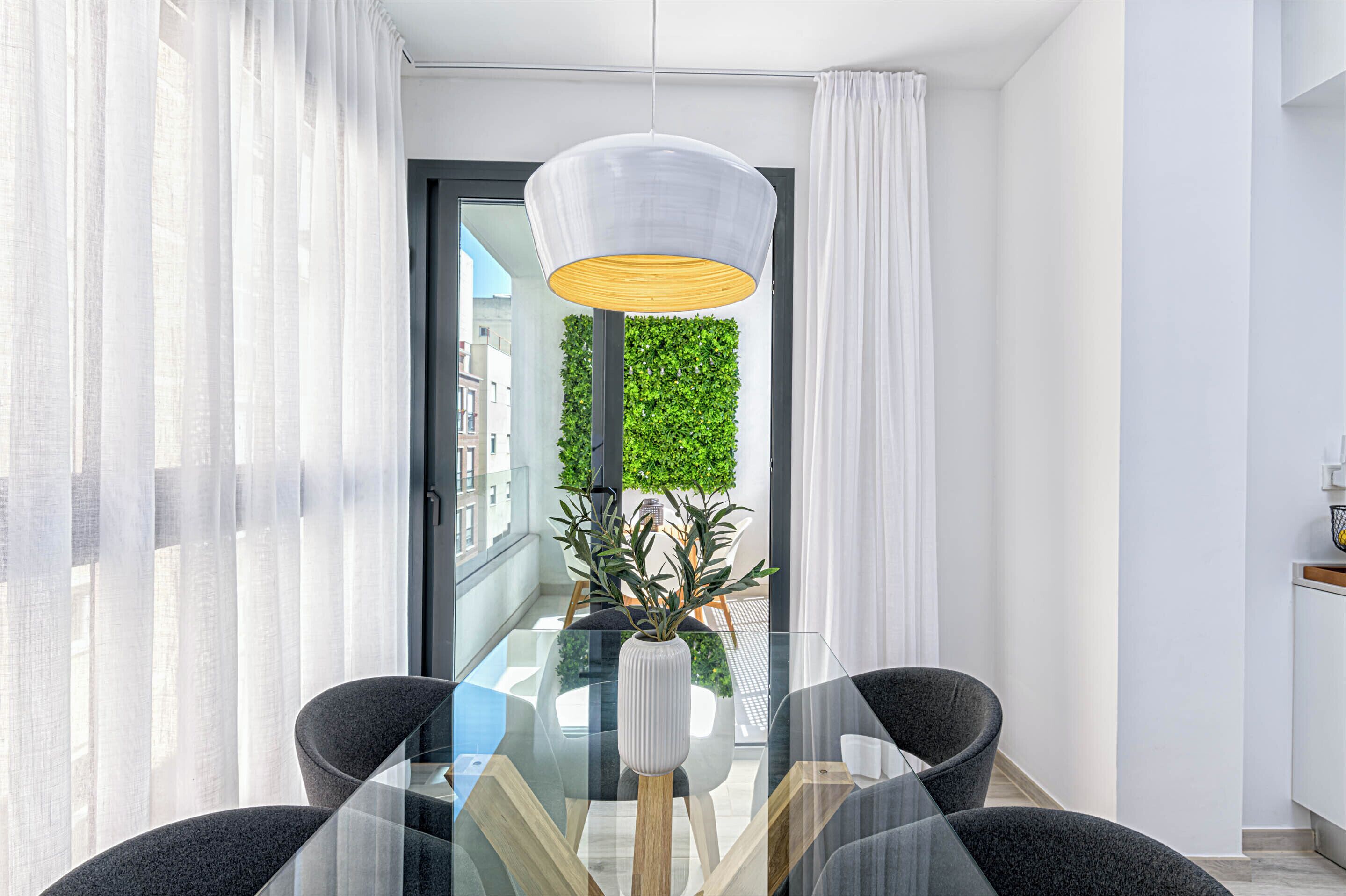 A modern dining area with a glass table, two chairs, a white vase with plants, and a hanging light fixture.