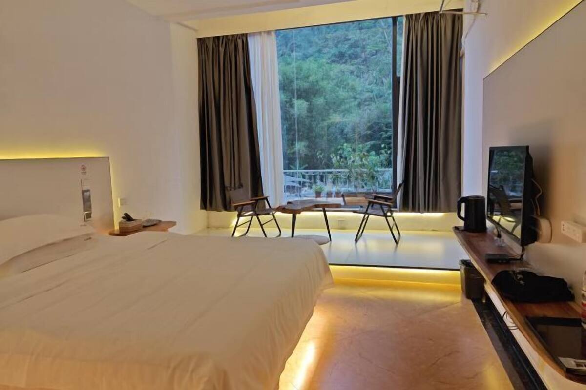 A hotel room with a bed, a desk with a chair, a television, and a view of greenery through the window.