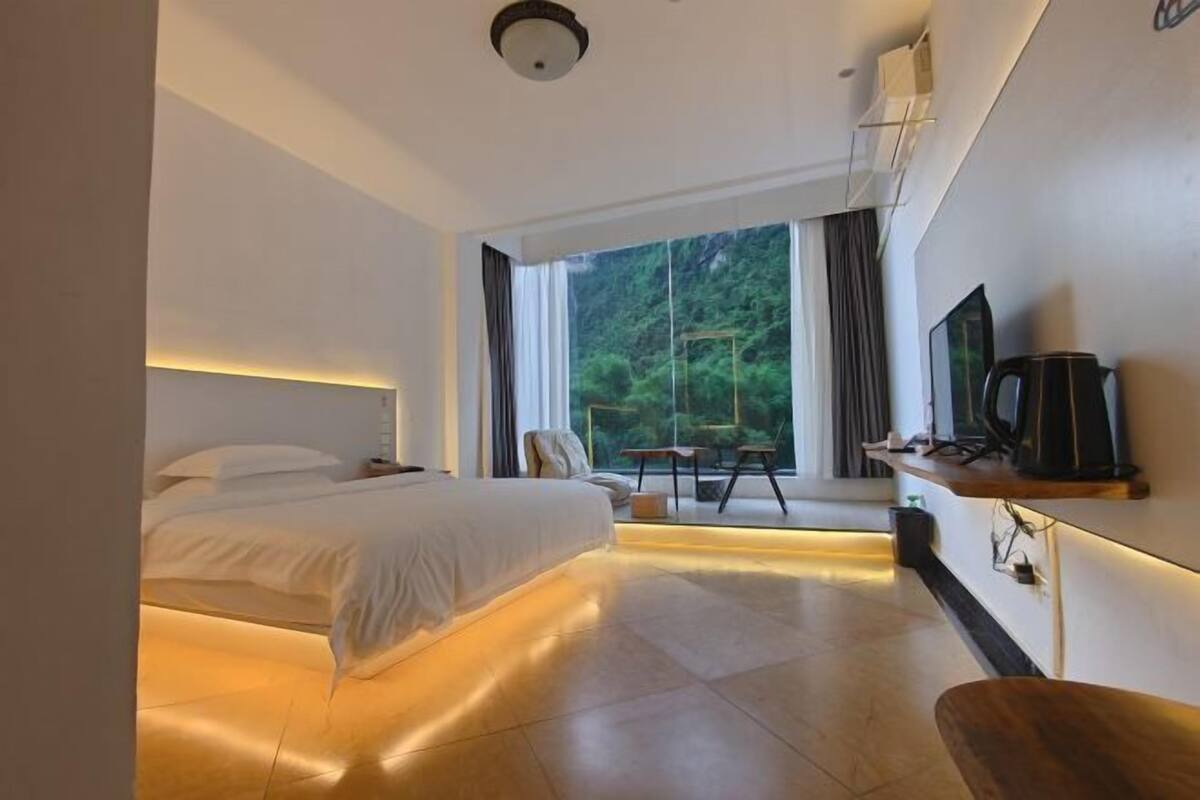 A modern hotel room with a bed, a desk, a chair, a TV, and a window with a view of trees.