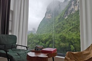 Ein Zimmer mit Blick auf eine gebirgige Landschaft, ein grüner Sessel, ein Holztischchen mit Teekanne und Buch sowie ein brauner Ohrensessel.
