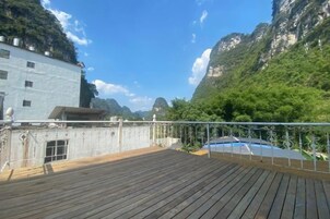 Una terraza de madera con baranda metálica, que da vista a un paisaje montañoso con vegetación y un edificio al fondo.