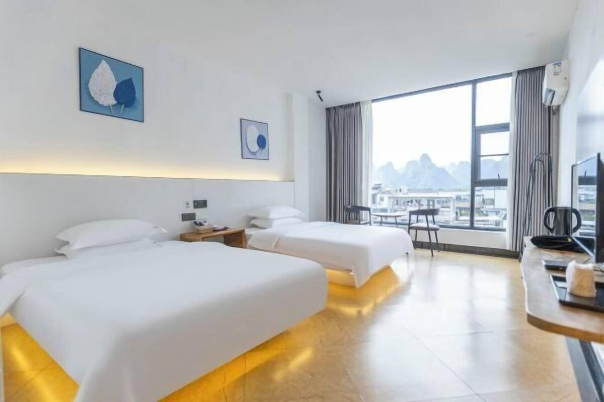 A hotel room with two beds, a desk, a chair, and a large window with a city view.