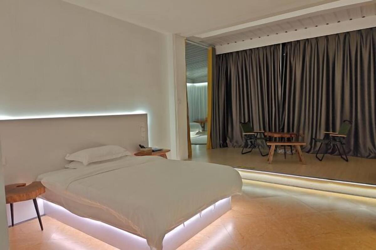 A modern bedroom with a large bed, bedside tables, a mirror, and a dining area with chairs and a table.