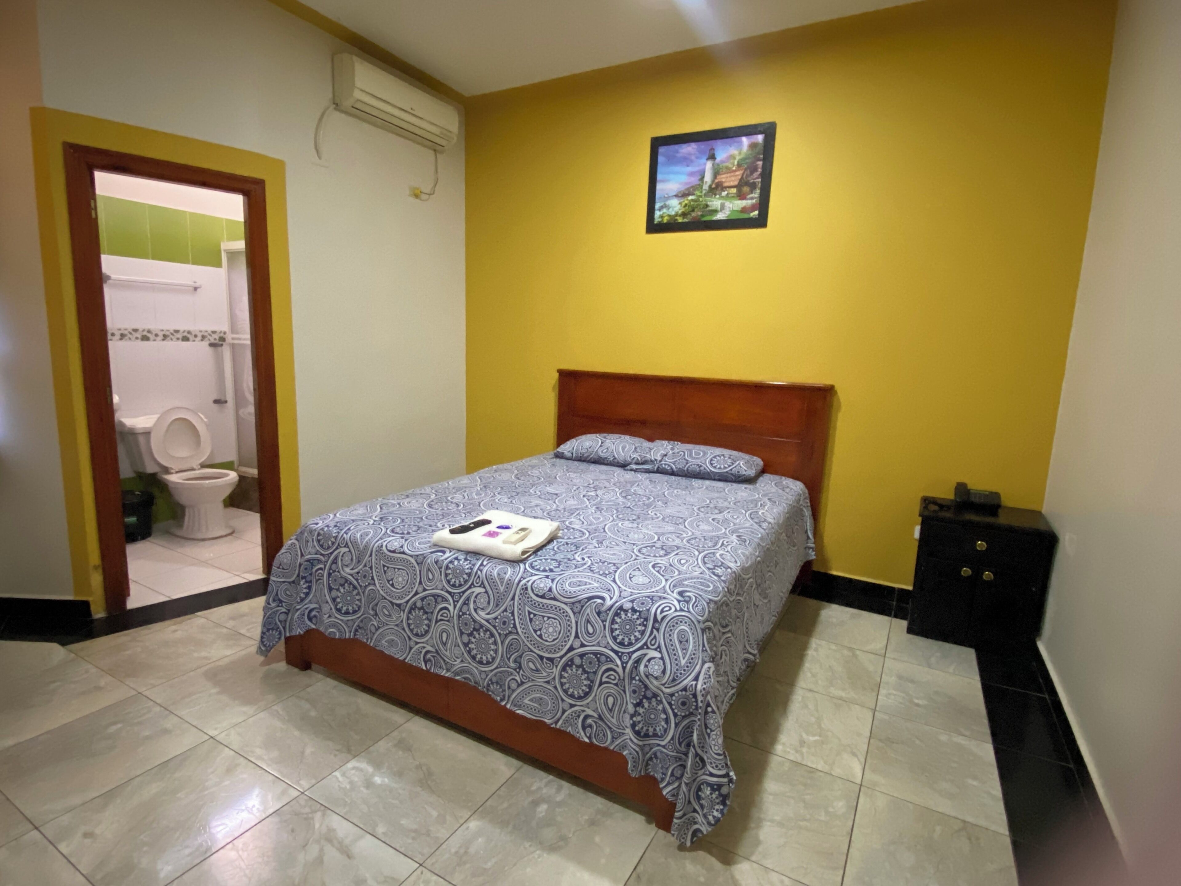A bedroom with a bed, a wooden headboard, a nightstand, a picture on the wall, and a bathroom visible through an open door.