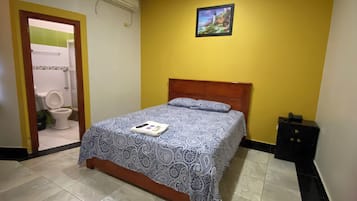 Ein Schlafzimmer mit Bett, Holz-Kopfteil, Nachttisch, einem Bild an der Wand und einem durchgehenden Türspalt sichtbaren Badezimmer.