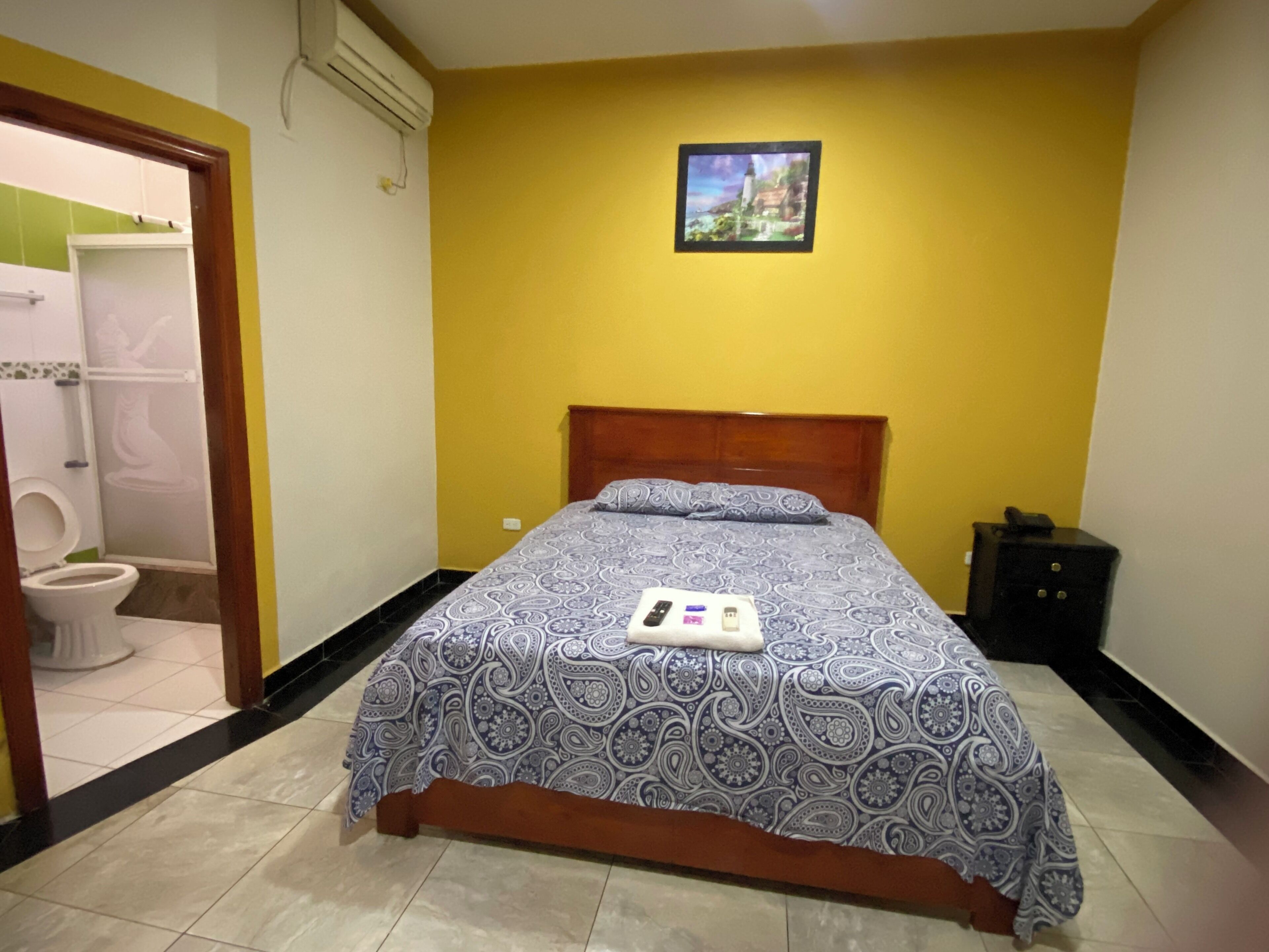 A bedroom with a bed, a bedside table, a picture on the wall, and a bathroom visible through an open door.
