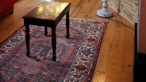 Una habitación con suelo de madera, un sofá rojo, un tapiz estampado, una mesa, una lámpara, una escalera y una pared de piedra.