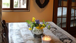 Una mesa de comedor con un centro de mesa floral y una vela encendida, ubicada en una habitación con vigas de madera y una ventana.