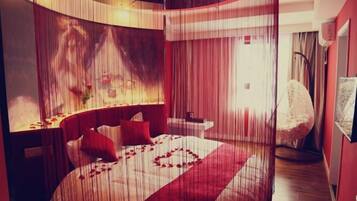 Un dormitorio moderno con una cama curvada, ropa de cama roja y una pantalla grande montada en la pared mostrando una escena romántica.