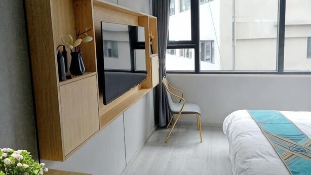 Et moderne hotelværelse med et ophængt fjernsyn, en seng med et dekorativt sengegærde, et vindue med udsigt til bygninger og en stol.