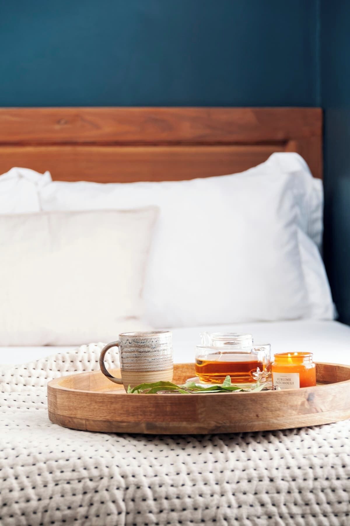 A wooden tray with a mug, a glass of amber liquid, and a candle labeled "Sublime Patchouli" on a bed with white pillows and a blue wall.