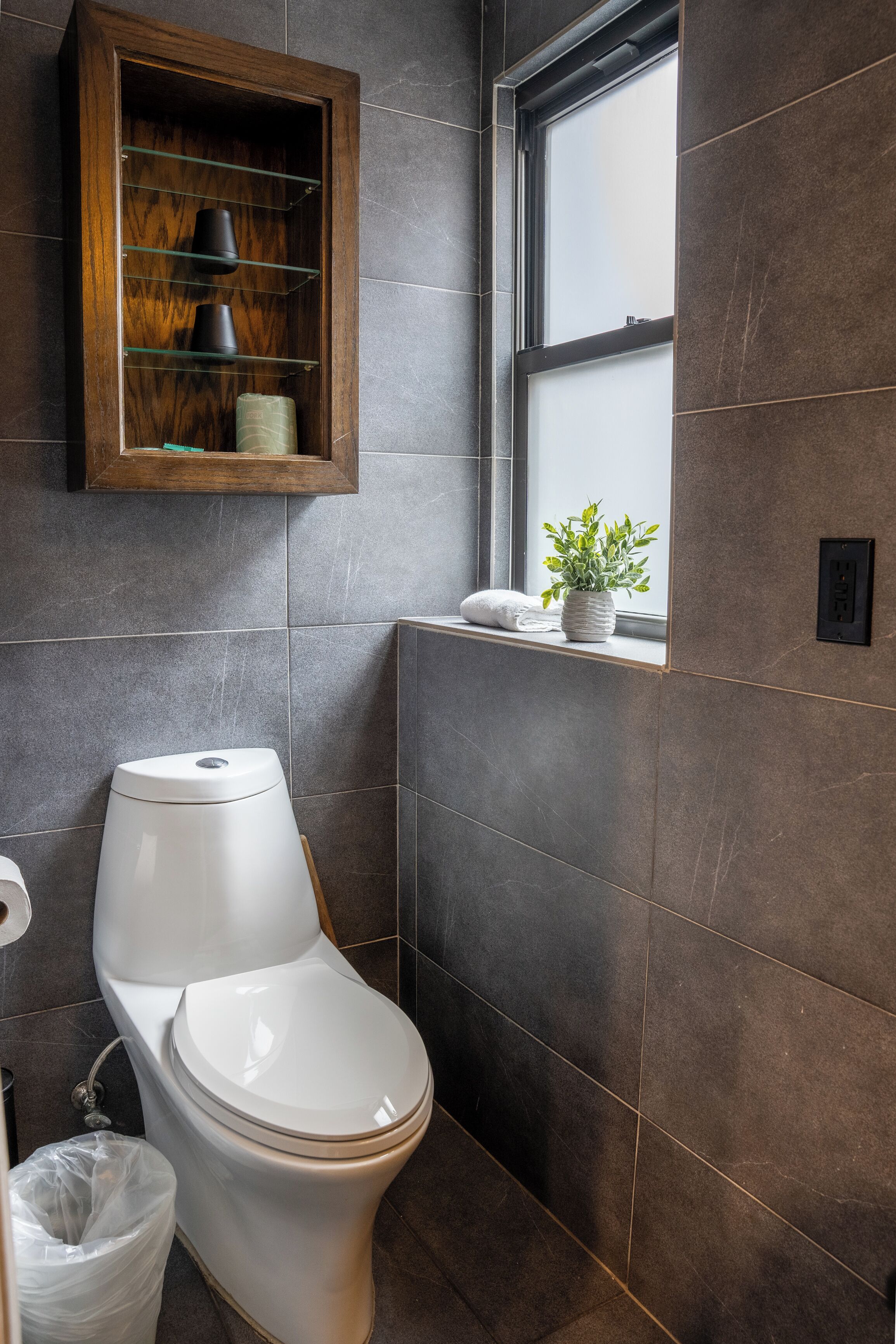 Un baño moderno con inodoro, un estante de vidrio con artículos de aseo y una ventana con planta.