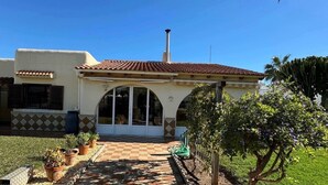 Una casa de dos plantas con techo de tejas, un porche cubierto con piso a cuadros y un jardín con diversas plantas y un cactus.