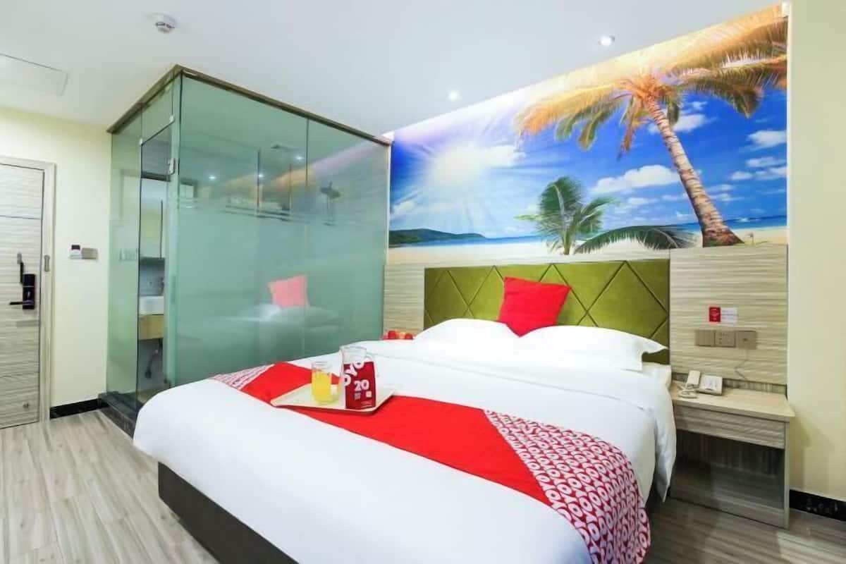 A hotel room with a bed, a glass partition, a mural of a beach scene, and a nightstand with a phone.