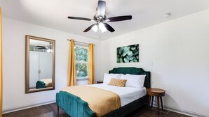 Una camera da letto con un letto, una ventola a soffitto, una finestra con tende, uno specchio e un quadro appeso al muro.