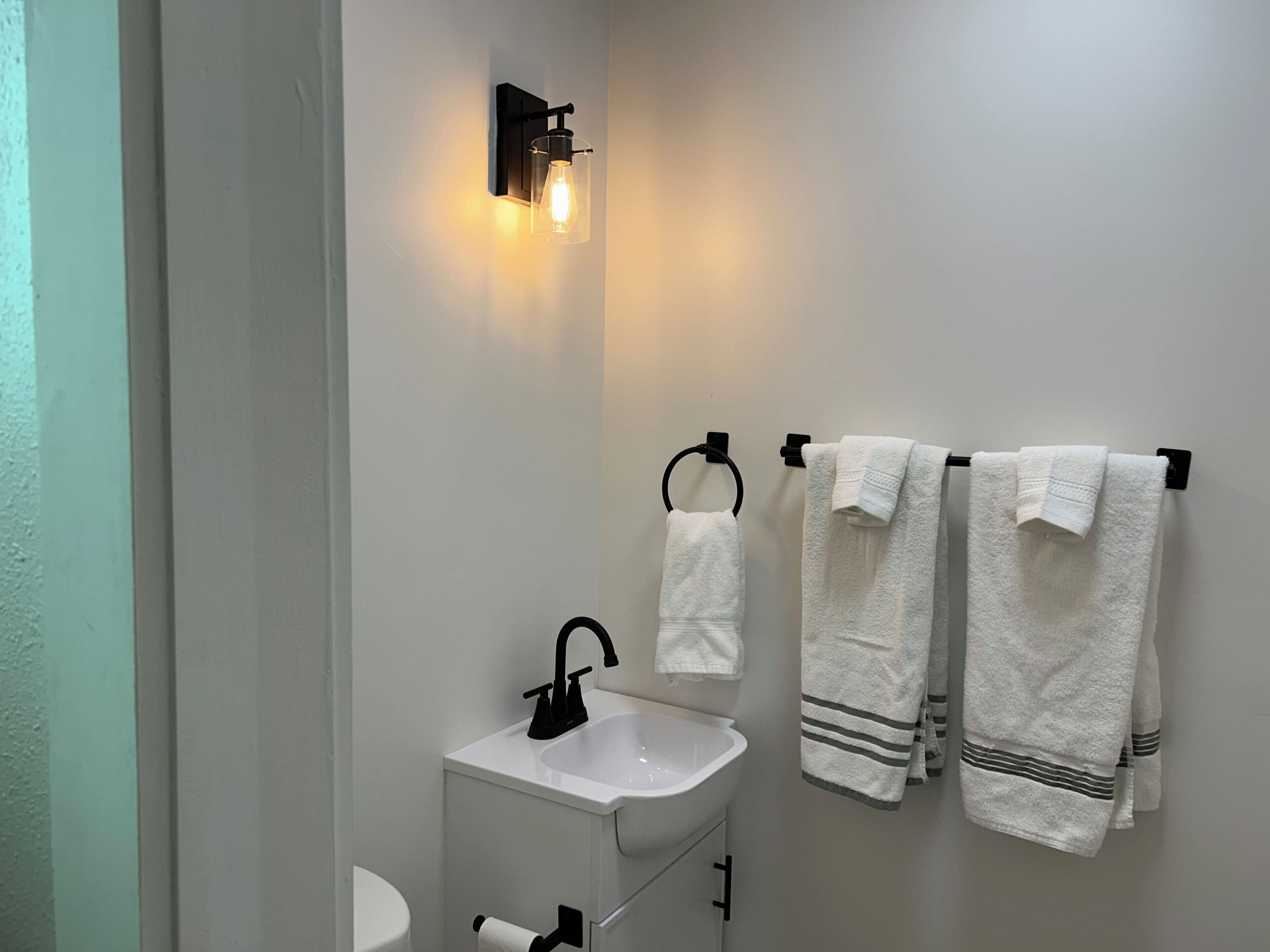 A bathroom with a white sink, black faucet, and a wall-mounted light fixture. There are three towels hanging on a towel rack.