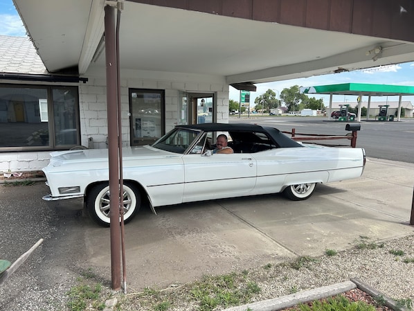 A white vintage car parked under a building with a gas station in the background.