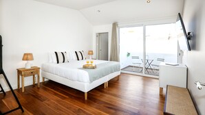 A modern bedroom with a large bed, wooden flooring, a bedside table with a lamp, a small table with a tray of oranges, and a TV on the wall. - Green Essence Duplex in Faro by Misha s Place (Faro)