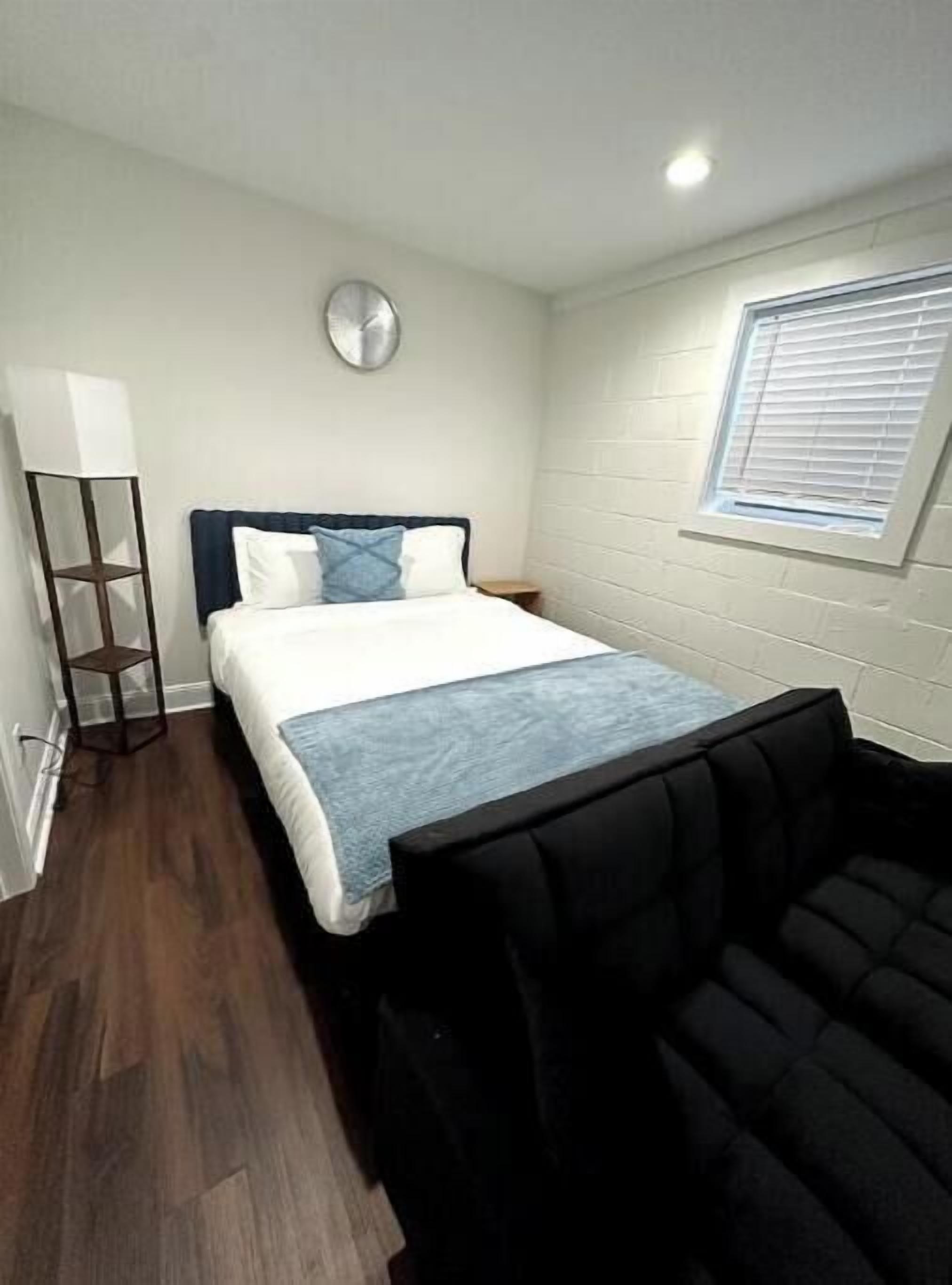 Una habitación pequeña y bien iluminada, con cama, mesita de noche, lámpara de pie y ventana con persianas.