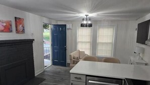 Et moderne køkken med mørkt bagpanel, en hvid bordplade og rustfrie stålapparater. Der er et spiseområde med et bord og stole, samt et stort vindue med persienner.