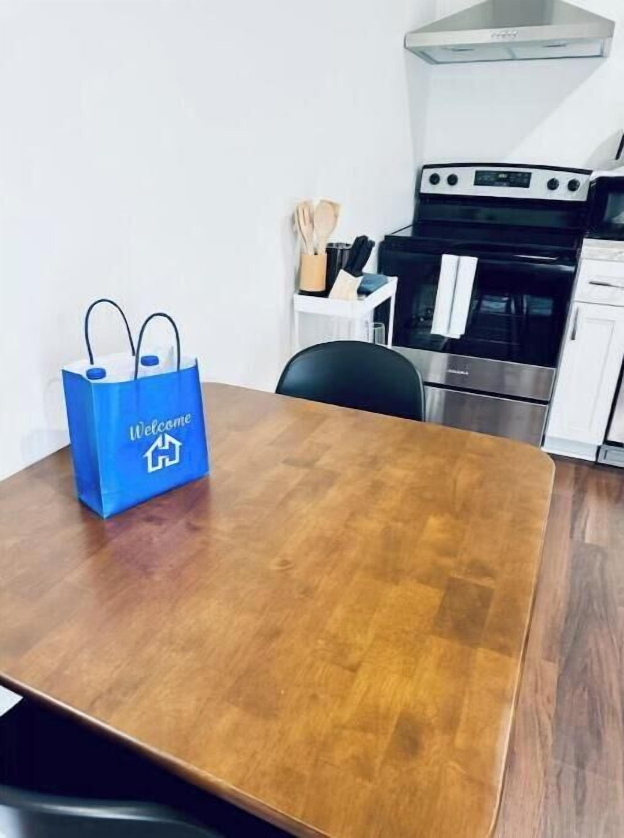 A wooden dining table with a blue welcome bag, a black chair, and a kitchen with a stove and oven in the background.