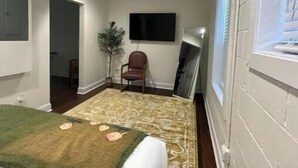 Ein schmales Hotelzimmer mit Bett, Wandfernseher, Stuhl und Fenster mit Jalousien.