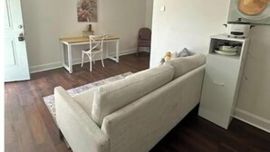 Ein Wohnzimmer mit einer Couch, einem Esstisch mit Stühlen, einer kleinen Küchenzeile mit Herd und einem Bild an der Wand.