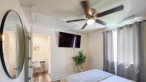 A hotel room with a bed, a ceiling fan, a TV, a plant, and a bathroom with a mirror.