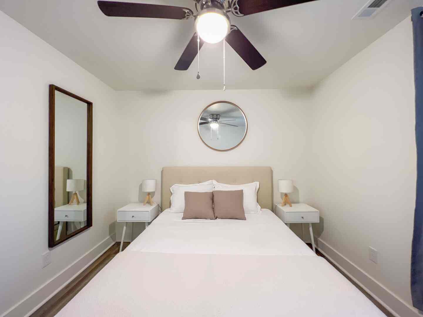 A hotel room with a bed, two bedside tables, a ceiling fan, a round mirror, and a wall-mounted light fixture.
