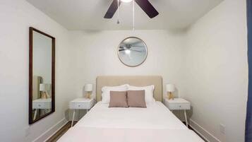 A hotel room with a bed, two bedside tables, a ceiling fan, a round mirror, and a wall-mounted light fixture.