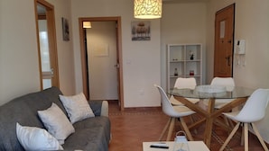 Una sala de estar con un sofá gris, una mesa de centro blanca y una zona de comedor con una mesa de vidrio y sillas blancas.