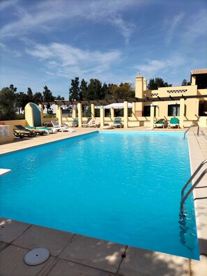 A swimming pool with lounge chairs and umbrellas, surrounded by a building with a tiled roof and a clear sky.