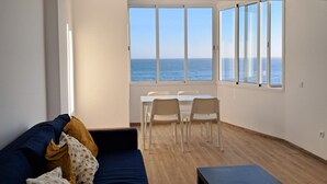 A modern living room with a large window overlooking the sea, a dark blue sofa, a small table, and a dining area with chairs. - T1 Pontemira 7C Vista Mar A C 80m Praia (Loulé)