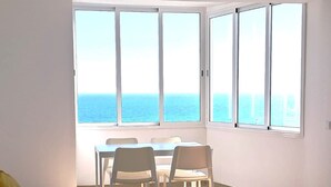 A modern room with a large window overlooking the sea, a blue sofa, a small table, and a dining area with chairs. - T1 Pontemira 7C Vista Mar A C 80m Praia (Loulé)
