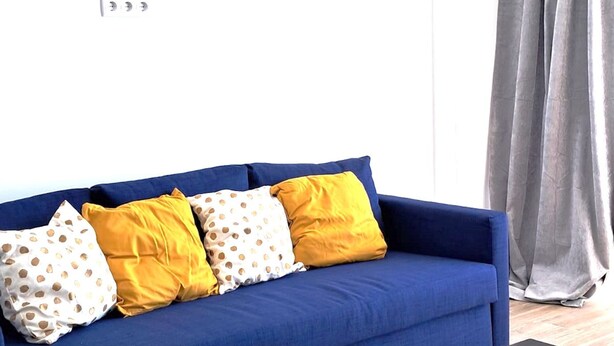 Ein blaues Sofa mit gelb-weißen Kissen, ein schwarzer Couchtisch und ein grauer Vorhang.