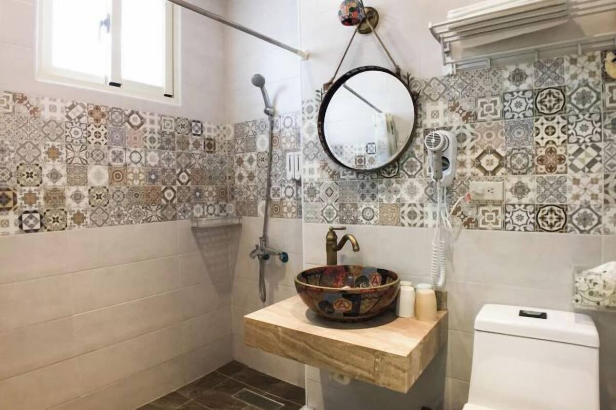 Ένα μπάνιο με πλακάκια στον τοίχο πίσω από τον νιπτήρα, έναν στρογγυλό καθρέφτη, έναν ξύλινο νιπτήρα, μια αποσπώμενη κεφαλή ντους και μια τουαλέτα.