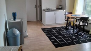 A compact kitchen with white cabinets, a sink, and a kettle. There's a dining area with a table and chairs, and a trash bin is visible. - Apartments in the heart of Hojer (Højer)
